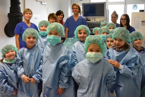 60 KInder erobern den OP (Krankenhaus Barmherzige Brüder Regensburg) Auf dem Foto sind die Mitarbeiterkinder zu sehen. Sie tragen Kittel und Mundschutz und grinsen frech in die Kamera.