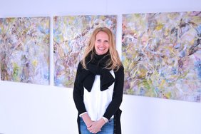 Ranftl Vernissage Erwachen (Krankenhaus Barmherzige Brüder Regensburg) Man sieht Doris Ranftl, eine blonde Frau mit mittellangen Haaren und schwarzem Schal, vor ihren Kunstwerken.