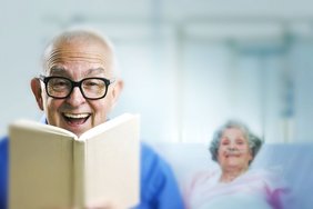 Dialog (Fotolia) Auf dem Bild ist ein älterer Mann mit Brille im Vordergrund, der lachend ein Buch liest. Im Hintergrund sieht man eine ältere Frau, die zufrieden lächelt.